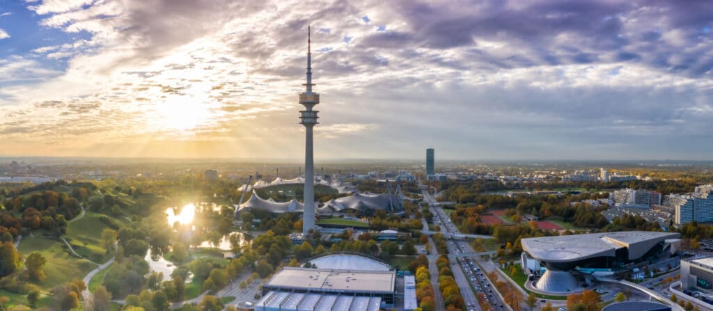 Panoramaaufnahme von München mit dem Olympiapark und dem markanten Olympiaturm im Vordergrund. Die ikonischen Zeltdachstrukturen des Olympiastadions erstrecken sich über das Gelände. Die Sonne geht hinter den Wolken unter und taucht die Szene in ein goldenes Licht. Im Hintergrund sind moderne Hochhäuser und Wohngebiete zu sehen, während die Straßen von Autos belebt sind.