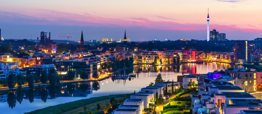 Abendliche Skyline von Dortmund mit Blick auf den Phoenix-See. Moderne Wohnhäuser mit beleuchteten Wegen im Vordergrund, während sich die Lichter der Stadt im ruhigen Wasser spiegeln. Der Florianturm und zwei Kirchtürme ragen in den farbenfrohen Sonnenuntergangshimmel, während im Hintergrund Industriebauten und Kräne zu sehen sind.