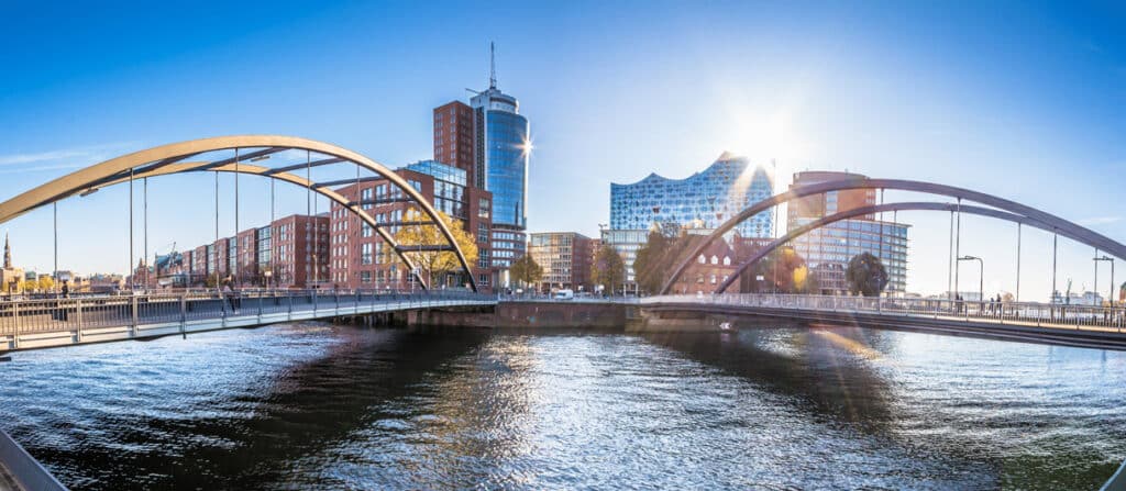 Panoramaansicht eines modernen Stadtviertels in Hamburg mit einer geschwungenen Brücke über das Wasser. Im Hintergrund sind moderne Hochhäuser und die berühmte Elbphilharmonie mit ihrer wellenförmigen Glasfassade zu sehen. Die Sonne strahlt hell am Himmel und wirft Lichtreflexionen auf das Wasser. Der blaue Himmel und die moderne Architektur spiegeln das lebendige Stadtbild wider.