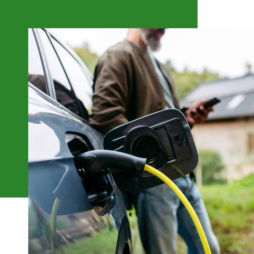 Ein Elektroauto wird mit einem gelben Ladekabel an einer Ladestation geladen. Im Hintergrund steht ein Mann mit Bart und legerer Kleidung, der auf sein Smartphone schaut. Im Hintergrund ist ein modernes Wohnhaus mit Solarpanelen auf dem Dach zu sehen, was auf nachhaltige Energiegewinnung hinweist. Die Szene vermittelt den Komfort des Ladens eines Elektrofahrzeugs zu Hause.