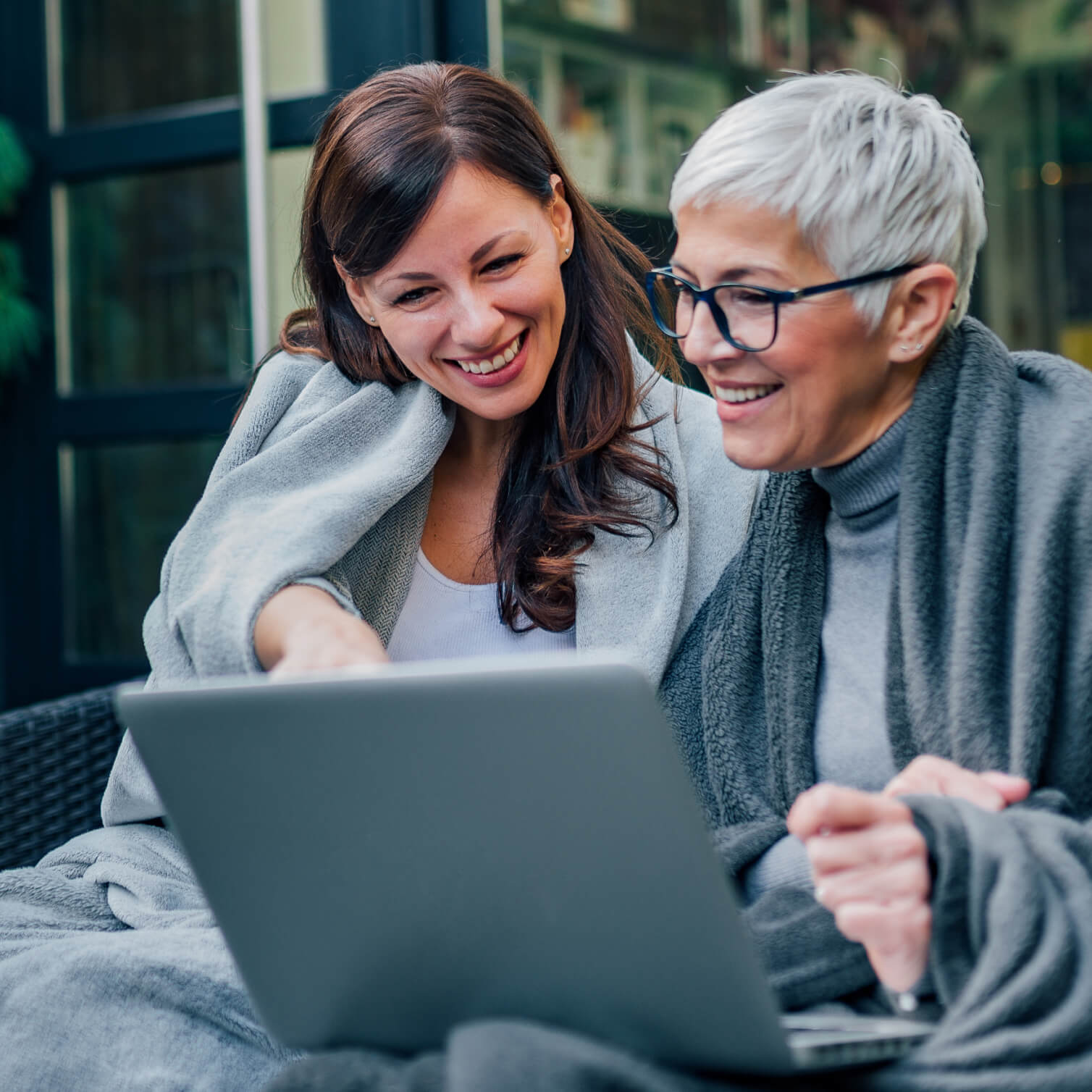 Zwei Frauen, eine jüngere und eine ältere mit grauen Haaren, sitzen gemeinsam draußen in warme Decken gehüllt und schauen lachend auf einen Laptop-Bildschirm. Sie wirken entspannt und zufrieden, während sie Informationen auf dem Laptop betrachten.