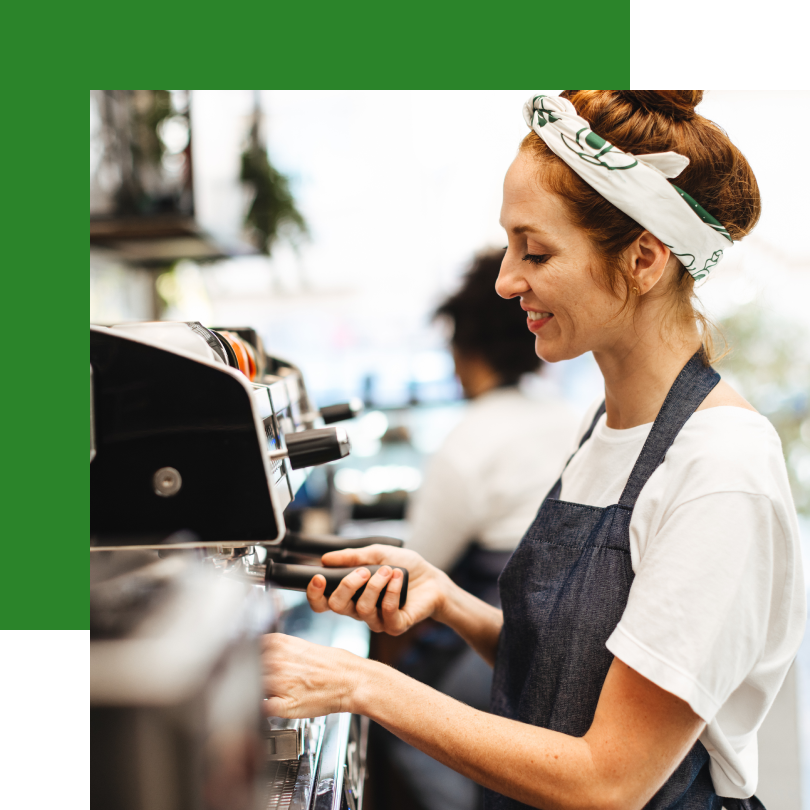 Eine lächelnde Barista mit hochgesteckten Haaren, einem weißen Kopftuch mit grünem Muster und einer blauen Schürze bereitet an einer Espressomaschine Kaffee zu. Sie hält einen Siebträger in der Hand und konzentriert sich auf ihre Arbeit. Im Hintergrund ist ein belebtes Café mit einer weiteren Mitarbeiterin und Regalen mit Kaffeebohnen und Tassen zu sehen.