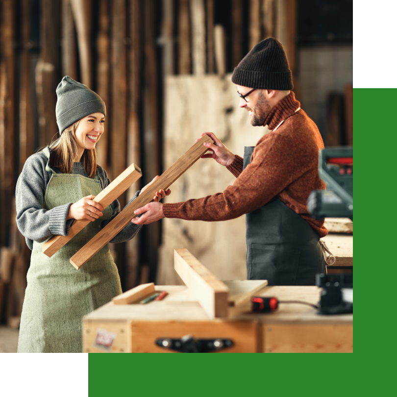 Ein Mann und eine Frau stehen in einer Werkstatt und arbeiten gemeinsam mit Holz. Beide tragen warme Kleidung, Schürzen und Mützen. Die Frau hält ein Holzbrett und lächelt, während der Mann ihr ein weiteres Brett reicht. Im Hintergrund sind Holzmaterialien und Werkzeuge sichtbar, was auf eine handwerkliche Umgebung hindeutet.